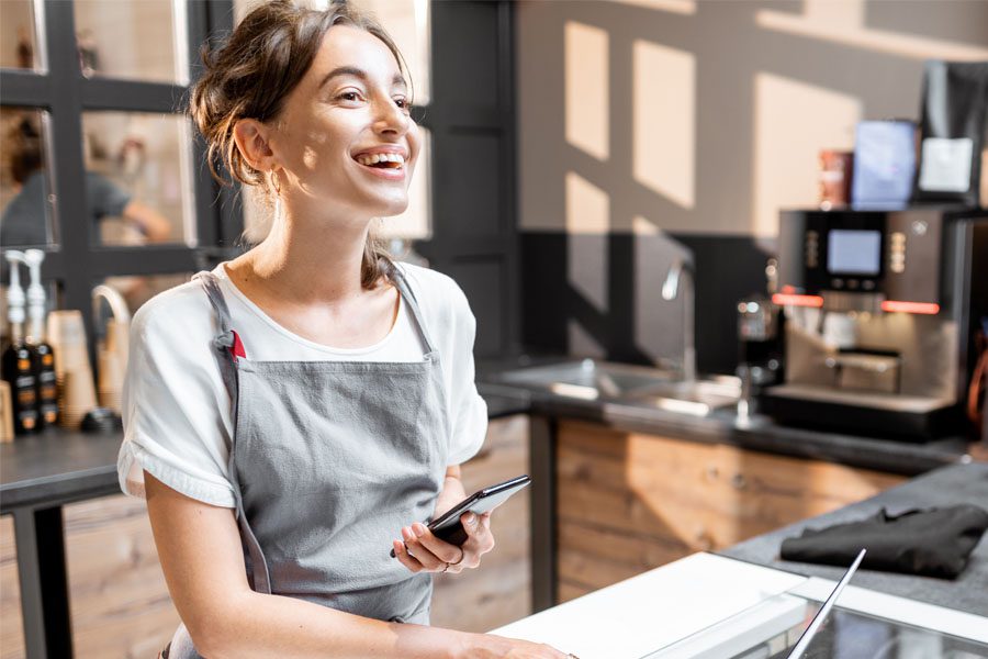 Business Insurance - Portrait of a Cheerful Female Small Business Owner Standing Behind the Counter of Her Ice Cream Shop and Using a Phone and Laptop