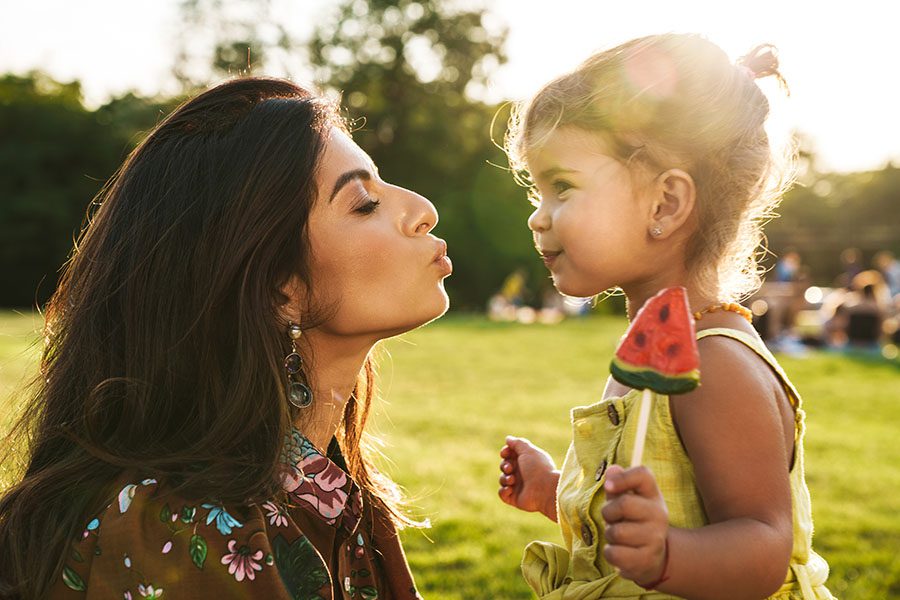 Contact - Mother Giving a Kiss to Her Young Daughter as She Eats a Piece of Watermelon in the Park